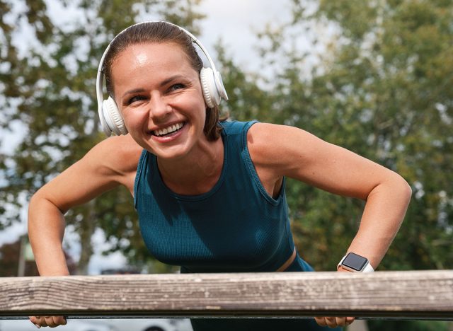 Young adult Caucasian woman in headphones performing push-ups on a bench during outdoor workout.